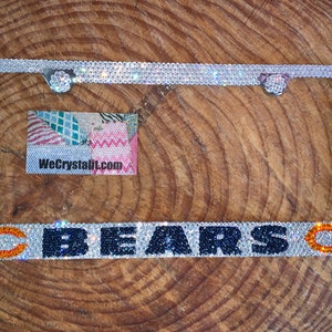 May include: A silver license plate frame with a clear crystal design. The frame has the word "BEARS" in black and blue crystals with a "C" on each side in orange and white crystals.