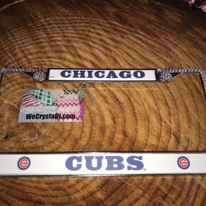 May include: A silver license plate frame with rhinestones around the border. The frame has two license plates, one that says "CHICAGO" in blue letters on a white background and another that says "CUBS" in blue letters on a white background with a red "C" logo on each side.
