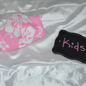 Pu&ograve; includere: Maschera facciale in tessuto floreale rosa e bianco con elastici bianchi. Un cartello nero con la parola "Kids" scritta in rosa.