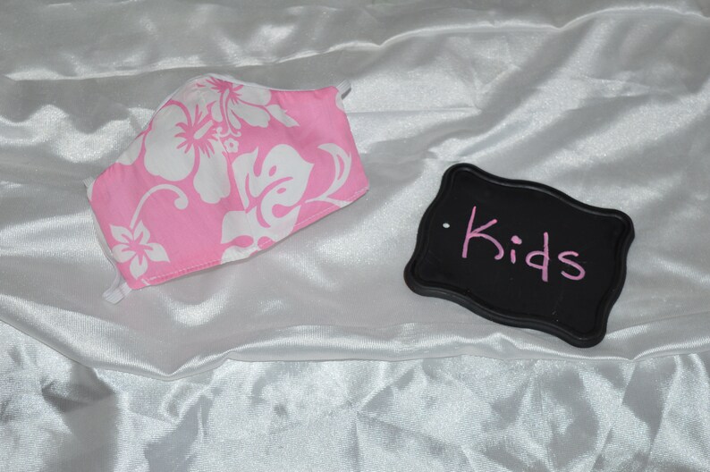 Pu&ograve; includere: Maschera facciale in tessuto floreale rosa e bianco con elastici bianchi. Un cartello nero con la parola "Kids" scritta in rosa.
