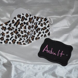 May include: A white face mask with a brown and black leopard print pattern. The mask is labeled "Adult" on a black sign.