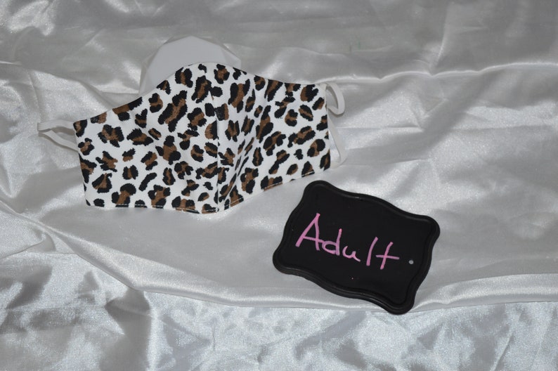 May include: A white face mask with a brown and black leopard print pattern. The mask is labeled "Adult" on a black sign.