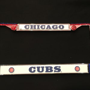 May include: License plate frame with red and blue rhinestones. The top bar reads "CHICAGO" in blue letters. The bottom bar reads "CUBS" with the team logo. The frame is a decorative accessory for a vehicle.
