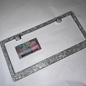 May include: A silver license plate frame with a border of clear rhinestones. The frame has two holes for screws on the top and bottom.