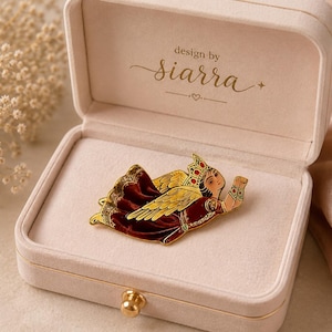 Pode incluir: Um broche dourado em forma de anjo, com detalhes em vermelho e amarelo, dentro de uma caixa de joias de veludo rosa. O broche apresenta um design detalhado de um anjo com coroa e asas abertas. A caixa aberta revela o broche e o texto "design by siarra".