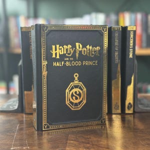 Könnte beinhalten: Ein schwarzes Buch mit goldenen Lettern und einem goldenen Design auf dem Einband. Der Titel lautet "Harry Potter und der Halbblutprinz".
