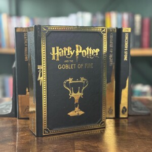Może przedstawiać: Czarna książka ze złotymi akcentami i tytułem "Harry Potter i Czara Ognia". Książka jest częścią zestawu książek o Harrym Potterze.