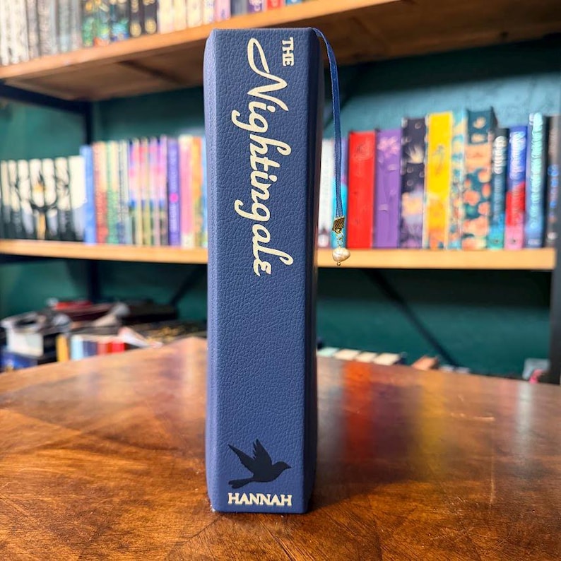 May include: A navy blue book titled "The Nightingales" with gold lettering. A silhouette of a bird is below the title, with the name "Hannah" at the bottom. A blue ribbon bookmark with a pearl charm is inserted.