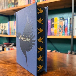 May include: A hardcover book titled "The Nightingale" by Kristin Hannah. The book has a blue cover with a silhouette of a bird and gold accents. The spine is black with gold bird designs.