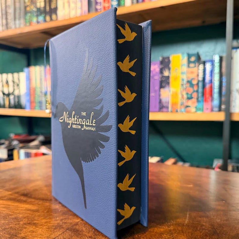 May include: A hardcover book titled "The Nightingale" by Kristin Hannah. The book has a blue cover with a silhouette of a bird and gold accents. The spine is black with gold bird designs.
