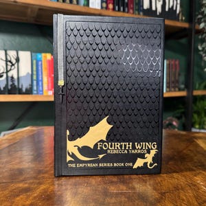 Op de afbeelding: Een zwart hardback boek met een gouden reliëf van een draak en de titel "Fourth Wing" van Rebecca Yarros. Het boek maakt deel uit van "The Empyrean Series" en is het eerste boek.
