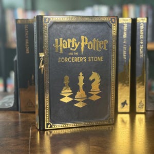 May include: A black hardback book with gold accents and the title "Harry Potter and the Sorcerer's Stone". The book is part of a set of books with gold spines.
