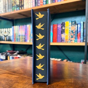 May include: A hardcover book with a navy blue spine featuring a repeating pattern of gold bird silhouettes. The book's pages are visible, and the background shows a bookshelf filled with various books, creating a literary theme.