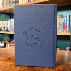 May include: A blue book with a textured cover. A black outline of the map of France is on the cover, with a heart symbol and a connecting line. The book is standing on a wooden surface.