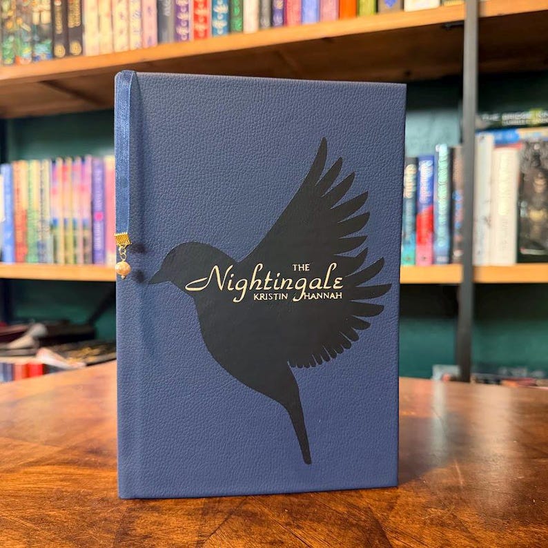 May include: A navy blue book titled "The Nightingale" by Kristin Hannah. The cover features a black silhouette of a bird with outstretched wings. A blue ribbon bookmark with a gold accent and a small bead hangs from the top.