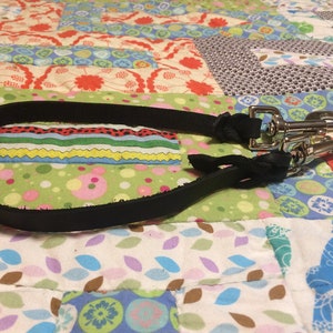 Puede incluir: Una correa de perro de cuero negro con dos clips de metal plateado. La correa está sobre una colcha de retazos de colores.