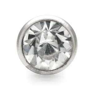 May include: A clear, faceted gemstone set in a silver bezel. The gemstone is round and has a high polish.