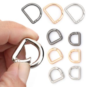 Könnte beinhalten: Eine Sammlung von D-Ring-Metallverschlüssen in verschiedenen metallischen Ausführungen, darunter Silber, Gold und Gunmetal. Die Verschlüsse werden in verschiedenen Größen gezeigt und sind für Taschen, Riemen und andere Bastelprojekte konzipiert.