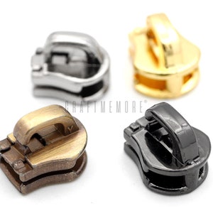 May include: Four zipper pulls in different colours: gold, silver, black, and bronze. The zipper pulls are all the same size and shape, and they have a small loop at the top.