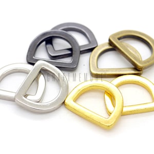 May include: A set of four D-rings in different finishes: silver, gold, antique brass, and black. The rings are all the same size and shape, with a rounded top and a flat bottom.