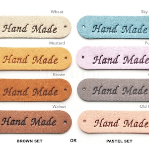 May include: Set of eight leather labels with the text "Hand Made" in a cursive font. The labels are in two colour sets: brown and pastel. The brown set includes wheat, mustard, brown, and walnut colours. The pastel set includes sky blue, purple, grey, and old rose colours.