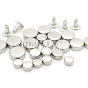 Könnte beinhalten: Eine Sammlung runder Nieten aus silbernem Metall mit glatter Oberfläche. Die Nieten sind auf einer weißen Oberfläche verstreut. CRAFT MEMORE TM ist auf der Oberfläche aufgedruckt.