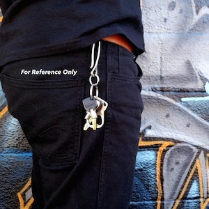 Pode incluir: Um chaveiro prateado com v&aacute;rias chaves presas, pendurado em um passador de cinto de uma pessoa. O chaveiro est&aacute; pendurado do lado direito do passador de cinto da pessoa. A pessoa est&aacute; vestindo jeans preto e uma camisa preta. O fundo &eacute; uma parede com grafite.