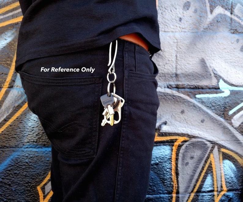 Pode incluir: Um chaveiro prateado com v&aacute;rias chaves presas, pendurado em um passador de cinto de uma pessoa. O chaveiro est&aacute; pendurado do lado direito do passador de cinto da pessoa. A pessoa est&aacute; vestindo jeans preto e uma camisa preta. O fundo &eacute; uma parede com grafite.