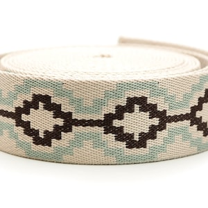 K&ouml;nnte beinhalten: Eine Rolle gewebten Stoffes mit einem geometrischen Muster. Der Stoff ist haupts&auml;chlich beige mit einem sich wiederholenden Design in Hellblau und Dunkelbraun. Das Muster zeigt Diamanten- und Zickzackformen. Die Rolle ist eng gewickelt.