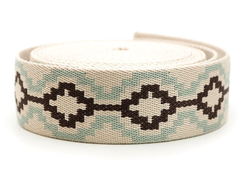 K&ouml;nnte beinhalten: Eine Rolle gewebten Stoffes mit einem geometrischen Muster. Der Stoff ist haupts&auml;chlich beige mit einem sich wiederholenden Design in Hellblau und Dunkelbraun. Das Muster zeigt Diamanten- und Zickzackformen. Die Rolle ist eng gewickelt.