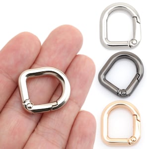 Puede incluir: Una mano sosteniendo un cierre de anilla en D plateado. Se muestran tres cierres adicionales: plateado, pavonado y dorado. Los cierres metálicos tienen una puerta con resorte para una fácil fijación a bolsos, correas u otros accesorios. Medidas en cm.
