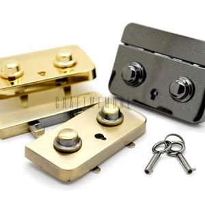 May include: Close-up of several metal purse clasps in gold and gunmetal finishes, with matching keys. The clasps have a rectangular shape with two rounded knobs and a keyhole. The keys are small and silver-coloured. The background is white.