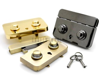 May include: Close-up of several metal purse clasps in gold and gunmetal finishes, with matching keys. The clasps have a rectangular shape with two rounded knobs and a keyhole. The keys are small and silver-colored. The background is white.