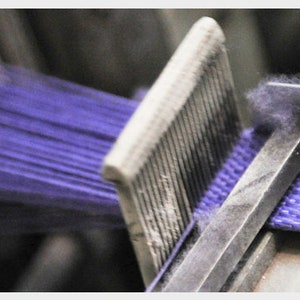 May include: Close-up of a purple woven strap being made on a machine. The strap is being pulled through a metal guide and then woven together.