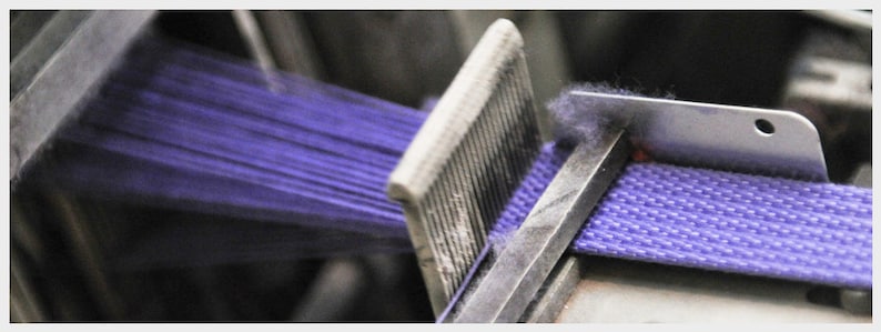 May include: Close-up of a purple woven strap being made on a machine. The strap is being pulled through a metal guide and then woven together.