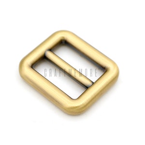 May include: A gold-toned metal buckle with a rectangular shape and a smooth finish. The buckle has two parallel slots for a strap or belt.