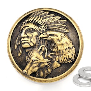Puede incluir: Un botón redondo de color latón con un diseño en relieve de un águila, un lobo y un indígena con tocado. El botón está rodeado por un anillo de metal plateado.