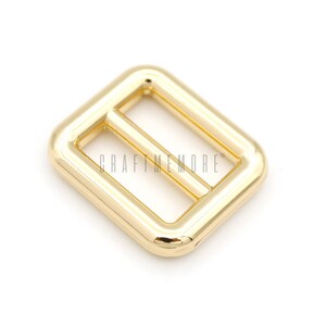 May include: Gold metal buckle with a rectangular shape and a smooth finish. The buckle has a center bar that is slightly raised.