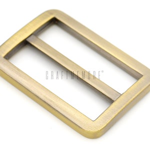 K&ouml;nnte beinhalten: Eine goldfarbene Metallschnalle mit rechteckiger Form und zwei parallelen Schlitzen. Die Schnalle ist f&uuml;r die Verwendung mit Riemen oder G&uuml;rteln konzipiert.