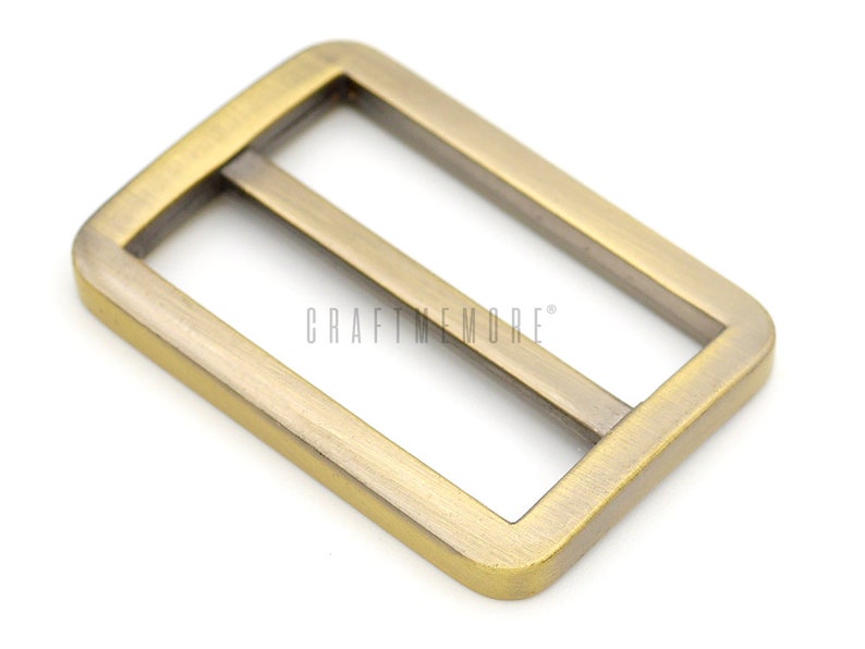 K&ouml;nnte beinhalten: Eine goldfarbene Metallschnalle mit rechteckiger Form und zwei parallelen Schlitzen. Die Schnalle ist f&uuml;r die Verwendung mit Riemen oder G&uuml;rteln konzipiert.