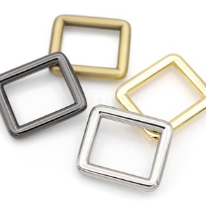 May include: Four square metal rings in different finishes: gold, silver, and black nickel. The rings are all the same size and shape.