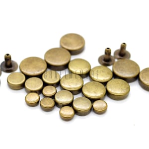 Peut inclure: Un assortiment de rivets et de clous de couleur laiton antique de différentes tailles. Les rivets ont un design à tige et à capuchon, tandis que les clous ont des dessus plats. Les articles sont présentés sur fond blanc, pour la maroquinerie ou la décoration vestimentaire.