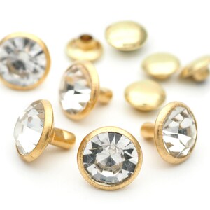 May include: A close-up of gold-toned metal studs with clear rhinestone accents. The studs have a flat back and a pointed top.