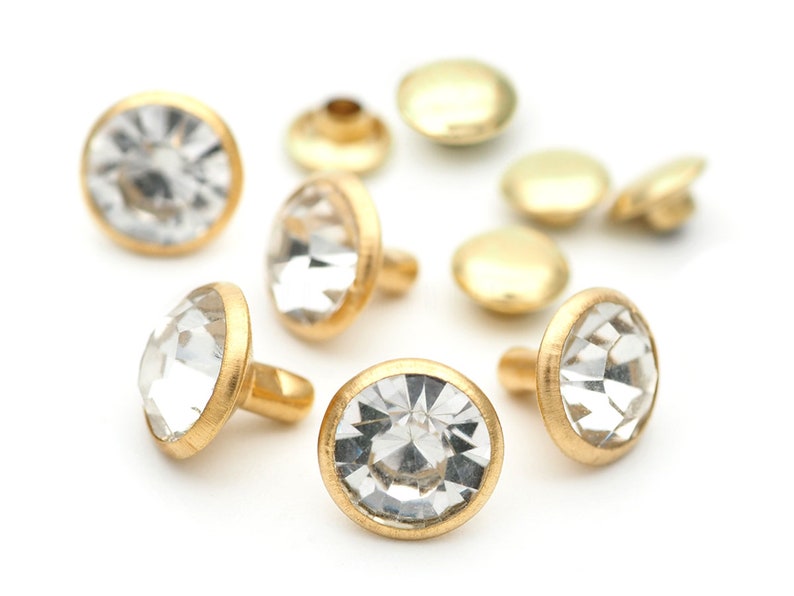 May include: A close-up of gold-toned metal studs with clear rhinestone accents. The studs have a flat back and a pointed top.