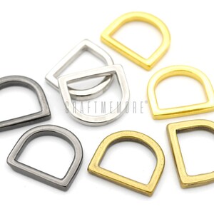 May include: Assortment of metal D-rings in gold, silver, and black finishes. The rings are teardrop shaped with a rounded opening.