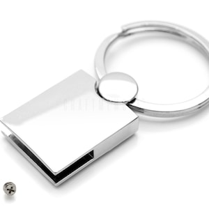 Puede incluir: Un llavero plateado con una etiqueta rectangular y un anillo redondo. Dos pequeños tornillos plateados son visibles en la superficie blanca.