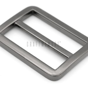 K&ouml;nnte beinhalten: Eine silberfarbene Metallschnalle mit rechteckiger Form und glatter Oberfl&auml;che. Die Schnalle hat eine Mittelstange, die durch die Schlaufe gleitet.