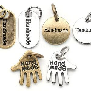 Könnte beinhalten: Sortiment aus sechs Metall-Anhängern mit der Gravur "Handmade". Die Anhänger sind in zwei Ausführungen: Antikmessing und Silber. Zwei Anhänger sind oval, zwei rund und zwei handförmig.
