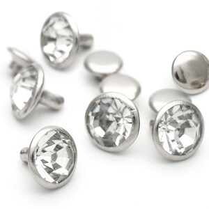 May include: A group of clear crystal rhinestones with silver settings. The rhinestones are arranged in a scattered pattern on a white background.