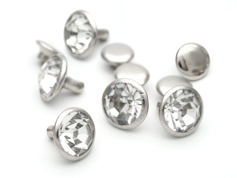 May include: A group of clear crystal rhinestones with silver settings. The rhinestones are arranged in a scattered pattern on a white background.